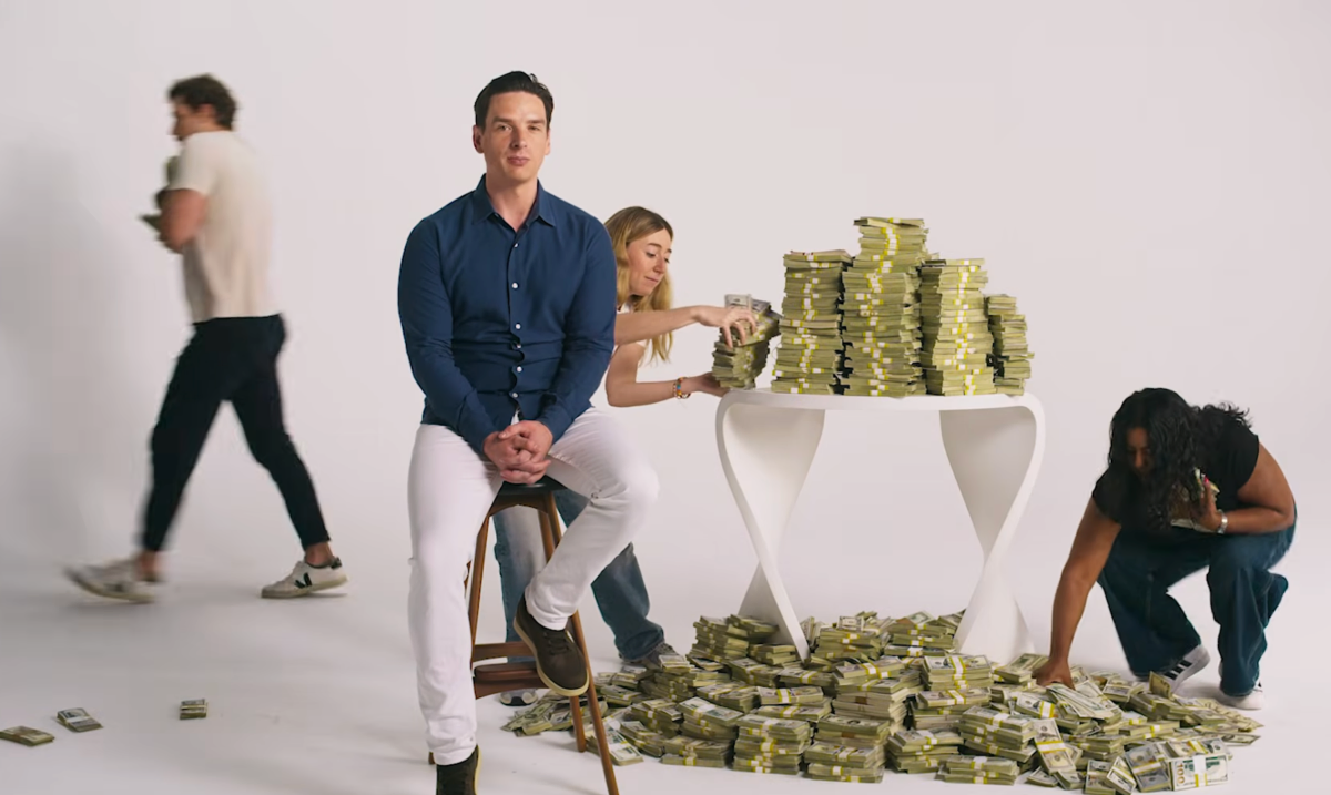 Man sitting on stool, two people stacking and collecting bundles of cash from table and floor, another person walking by.