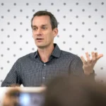 Man in checkered shirt speaking in front of a dotted backdrop, gesturing with one hand raised.