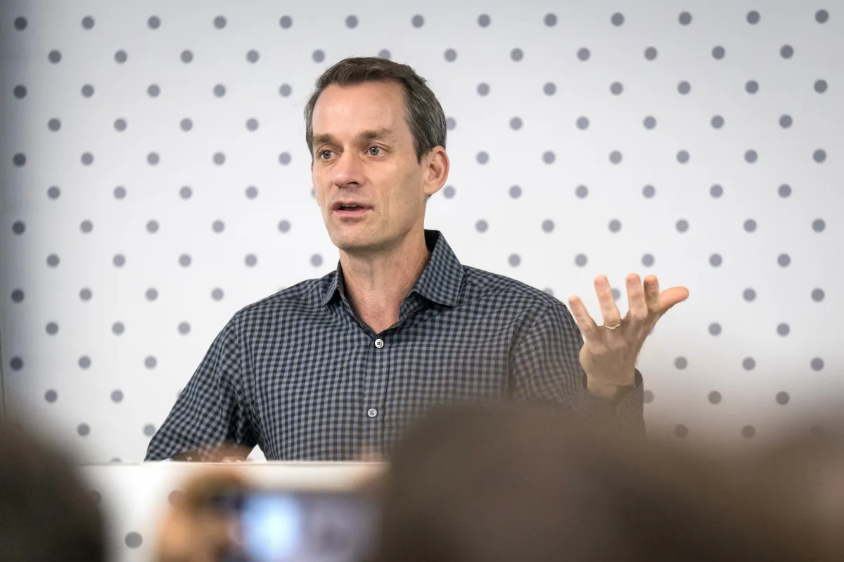Man in checkered shirt speaking in front of a dotted backdrop, gesturing with one hand raised.