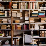 Bookshelves filled with various books and papers, a step ladder in front, and small labels with handwritten text on the sh...