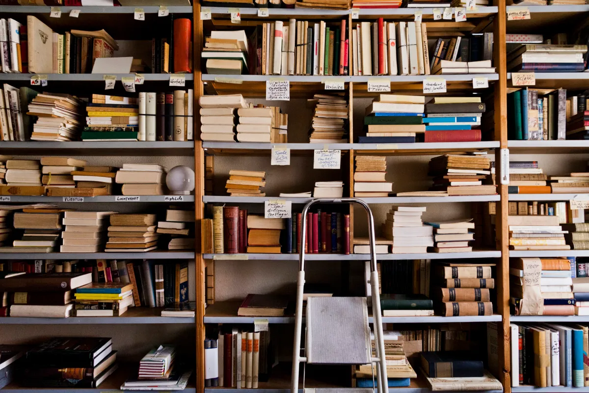 Bookshelves filled with various books and papers, a step ladder in front, and small labels with handwritten text on the sh...