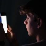 Young person in a dark room looking at a bright smartphone screen, face illuminated by the light.
