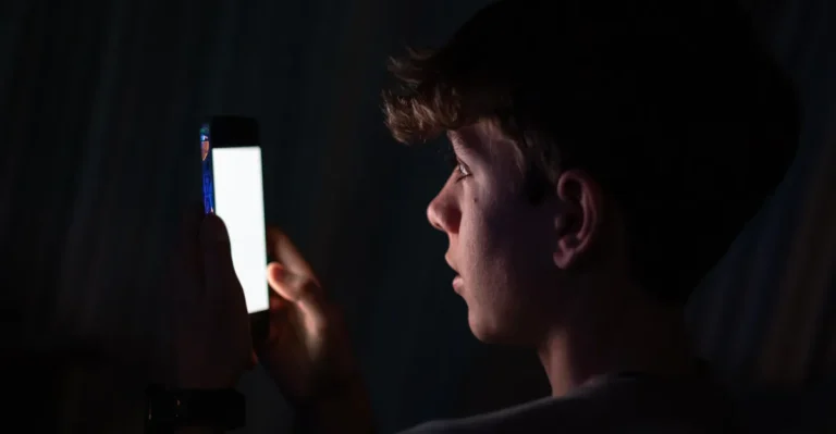 Young person in a dark room looking at a bright smartphone screen, face illuminated by the light.
