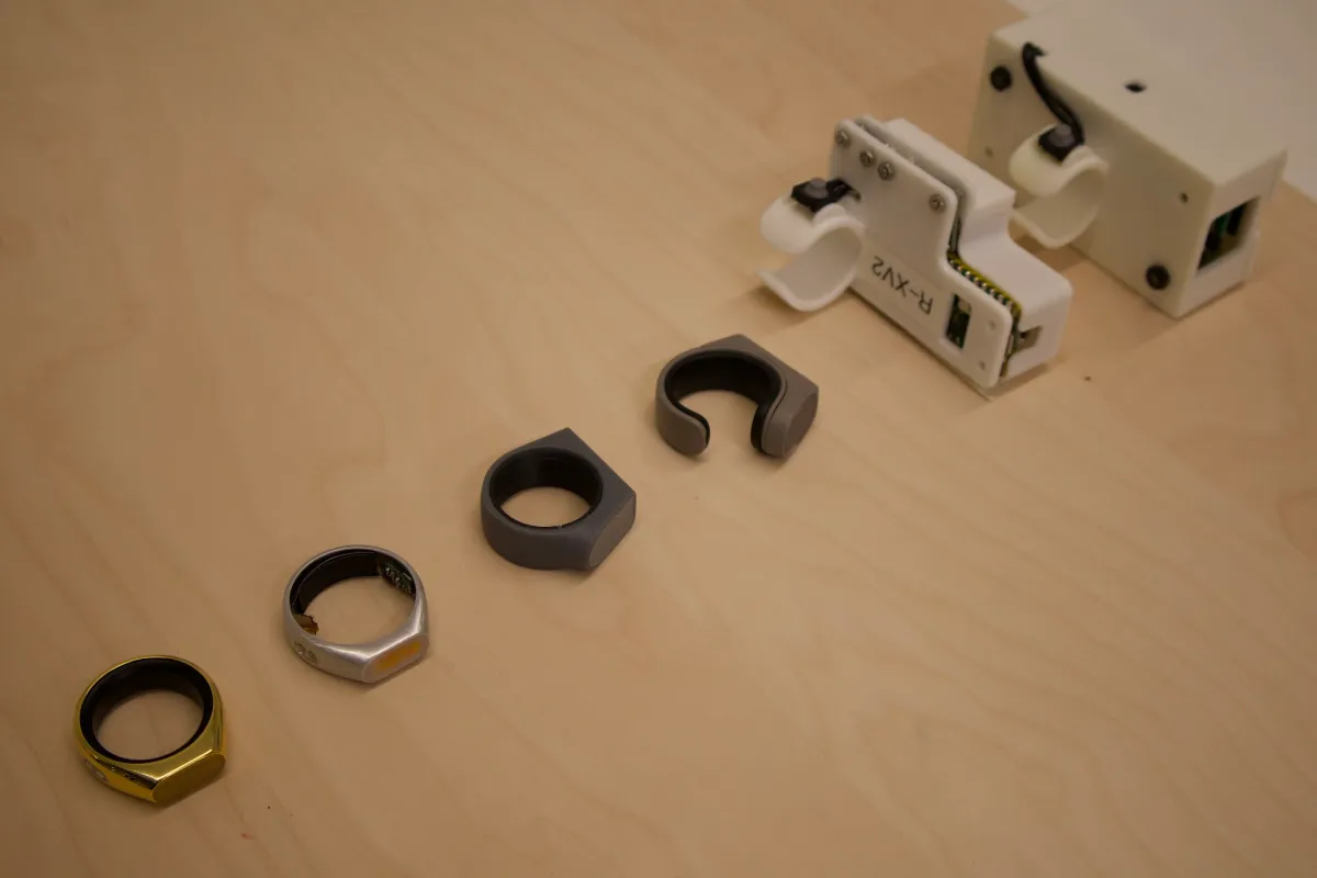 Three rings in different finishes and a white electronic device on a wooden surface.