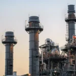 Industrial gas-fired power plant with multiple tall chimneys and complex metal structures under a clear sky.