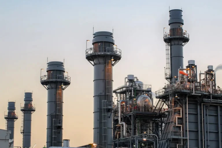 Industrial gas-fired power plant with multiple tall chimneys and complex metal structures under a clear sky.
