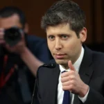 Man in a suit giving a thumbs-up at a microphone, with a photographer in the background capturing the moment.