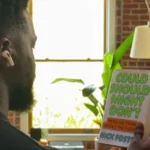 Person with earbuds holds a book titled "Could Should Might Don't" in a room with brick walls, a lamp, and a plant.