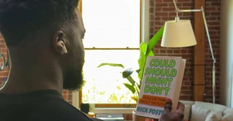 Person with earbuds holds a book titled "Could Should Might Don't" in a room with brick walls, a lamp, and a plant.