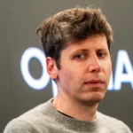 Person with short hair in a gray sweater standing in front of a blurred background with partial text "AI" visible.