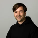 Smiling person with short dark hair wearing a black hoodie against a plain gray background.
