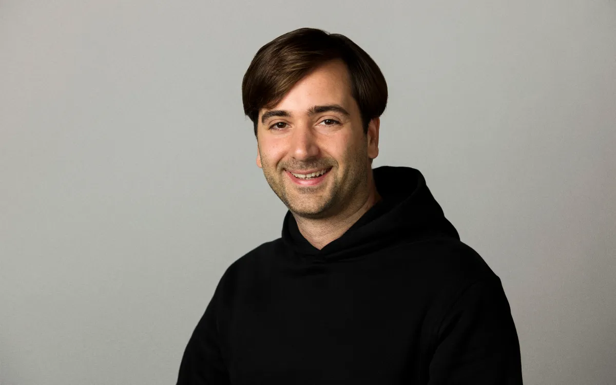 Smiling person with short dark hair wearing a black hoodie against a plain gray background.