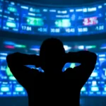 Silhouette of a person with hands on head, looking at large, brightly lit stock market screens displaying numbers and graphs.