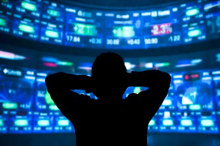Silhouette of a person with hands on head, looking at large, brightly lit stock market screens displaying numbers and graphs.