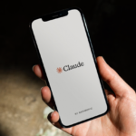 Hand holding a smartphone displaying the Claude app logo with "by Anthropic" text on a simple background.