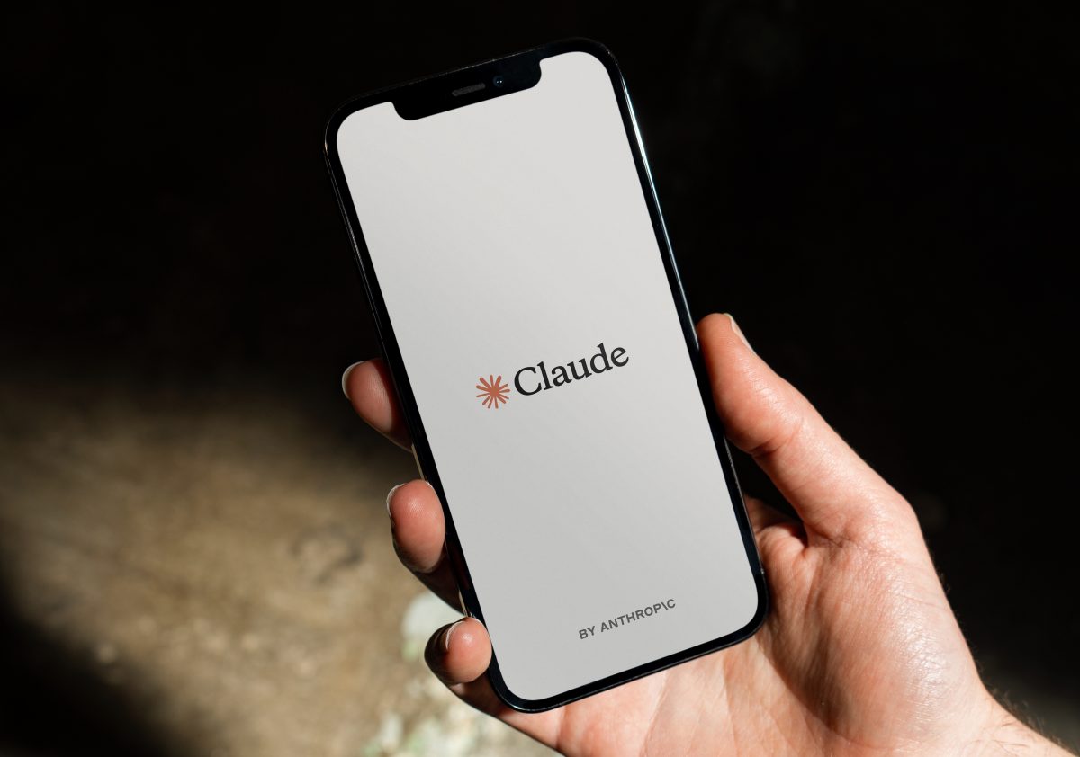 Hand holding a smartphone displaying the Claude app logo with "by Anthropic" text on a simple background.