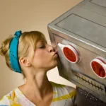 A woman kisses a large robot head with red eyes, wearing a blue headband and colorful shirt, against a plain background.