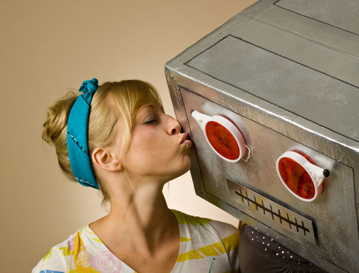 A woman kisses a large robot head with red eyes, wearing a blue headband and colorful shirt, against a plain background.