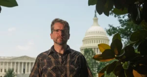 Man in glasses and plaid shirt stands in front of the US Capitol building, framed by green leaves.