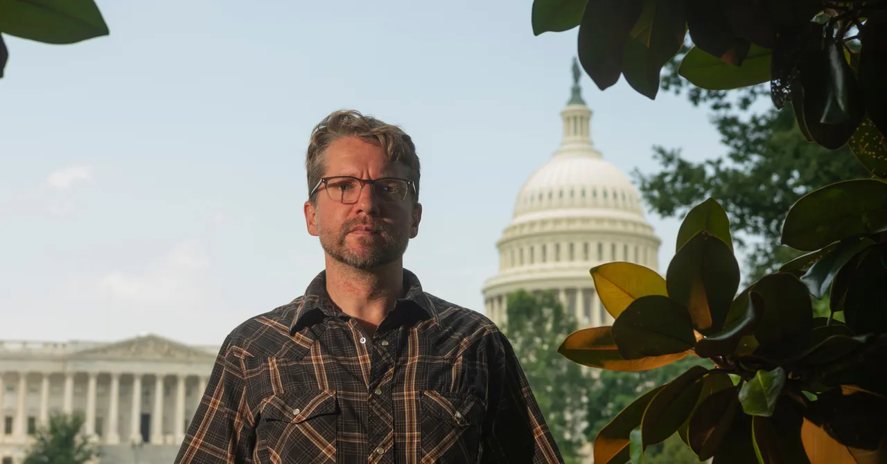 Man in glasses and plaid shirt stands in front of the US Capitol building, framed by green leaves.