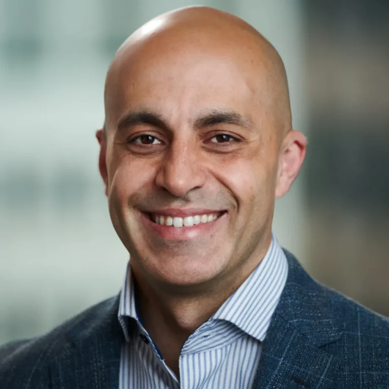 Smiling bald man in a gray suit jacket and striped shirt, with a blurred background.