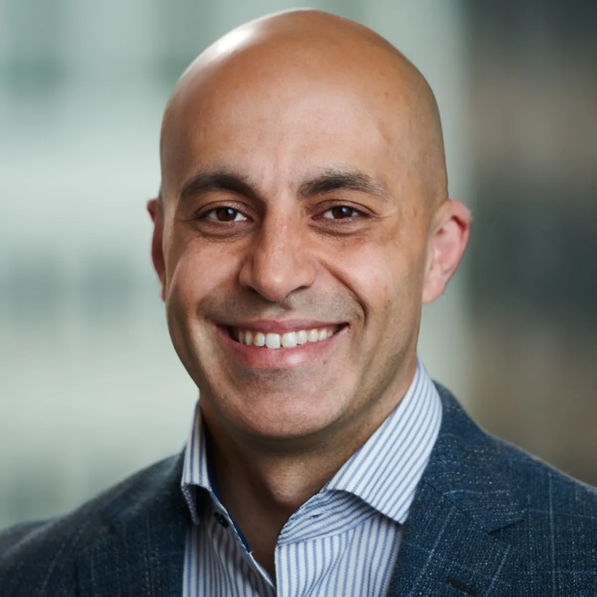 Smiling bald man in a gray suit jacket and striped shirt, with a blurred background.