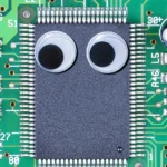 Microchip on a green circuit board with two googly eyes attached, surrounded by labeled electronic components.