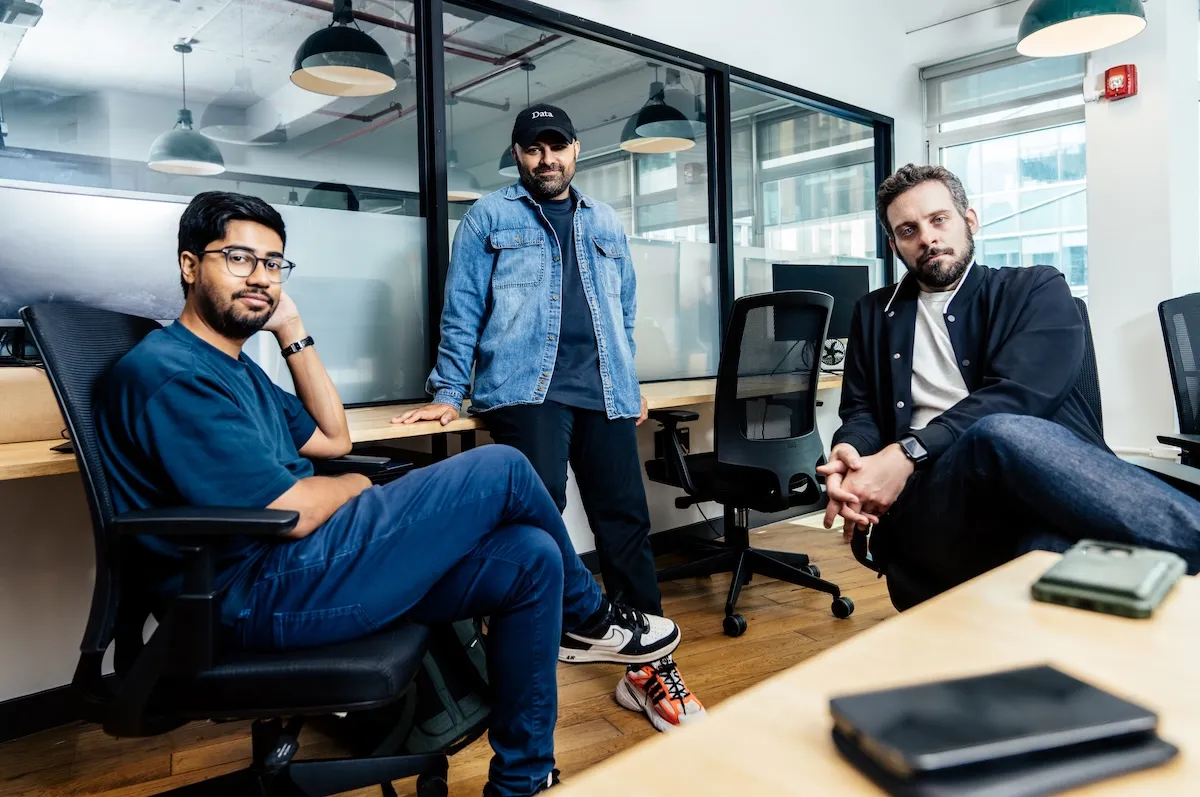 Three men in casual attire sit and stand in a modern office with large windows and hanging lights.