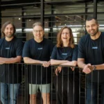 Four people in matching "June" shirts smiling, standing behind a railing in an office setting.