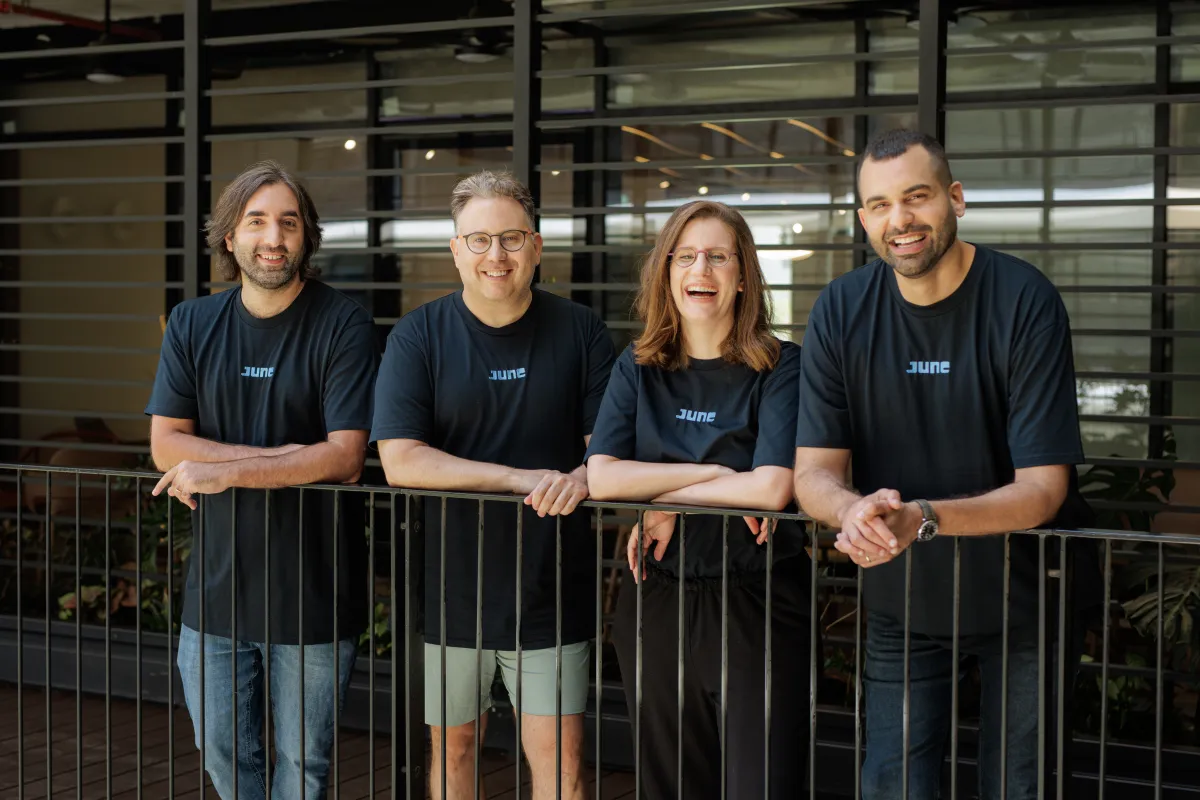 Four people in matching "June" shirts smiling, standing behind a railing in an office setting.