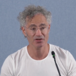 Man with curly hair and glasses speaking into a microphone, wearing a white shirt against a plain blue background.