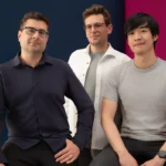 Three men sitting in front of a blue and pink background, wearing casual shirts, looking at the camera.