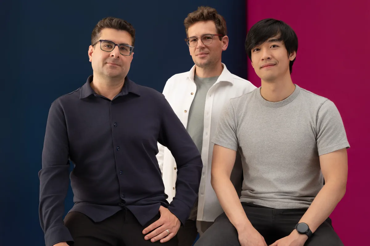 Three men sitting in front of a blue and pink background, wearing casual shirts, looking at the camera.