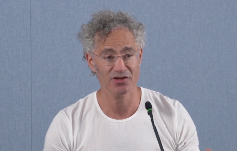 Man with curly hair and glasses speaking into a microphone, wearing a white shirt against a plain blue background.