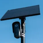 Black surveillance camera and solar panel mounted on a pole against a clear blue sky.