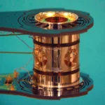 Shiny metallic cylindrical component with intricate design and wiring, set against a turquoise background.