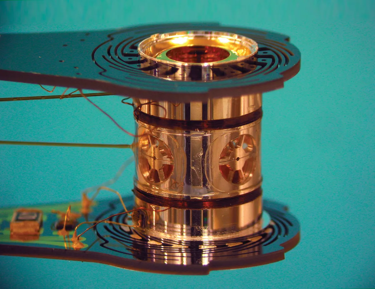 Shiny metallic cylindrical component with intricate design and wiring, set against a turquoise background.