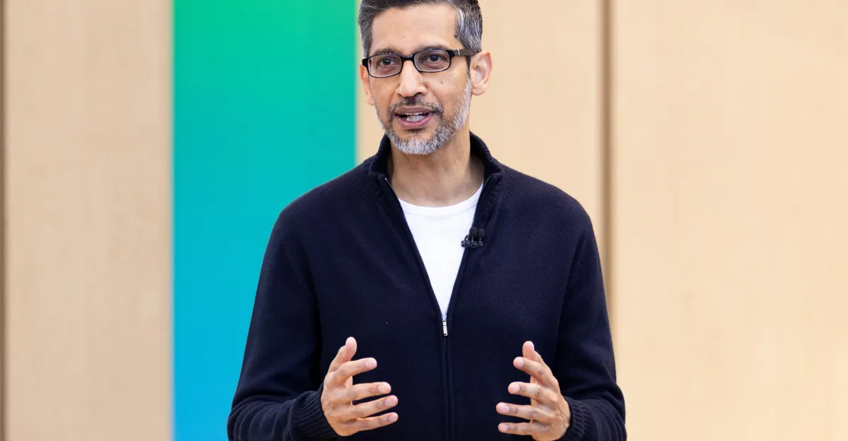 A man in a black zip-up sweater speaks with hand gestures in front of a green and beige background.