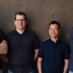 Four men standing against a textured wall, all wearing dark shirts, smiling at the camera.