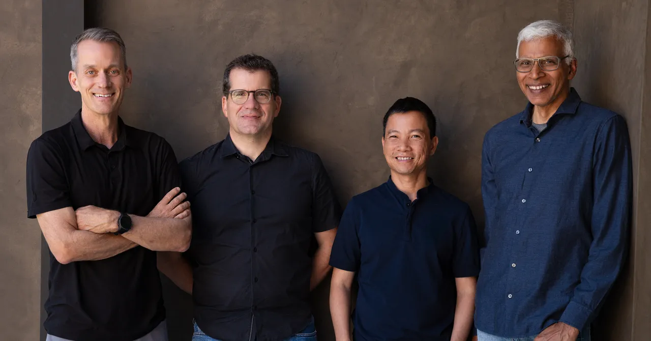 Four men standing against a textured wall, all wearing dark shirts, smiling at the camera.