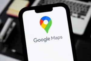 Smartphone displaying Google Maps logo and text, with a blurred laptop keyboard in the background.