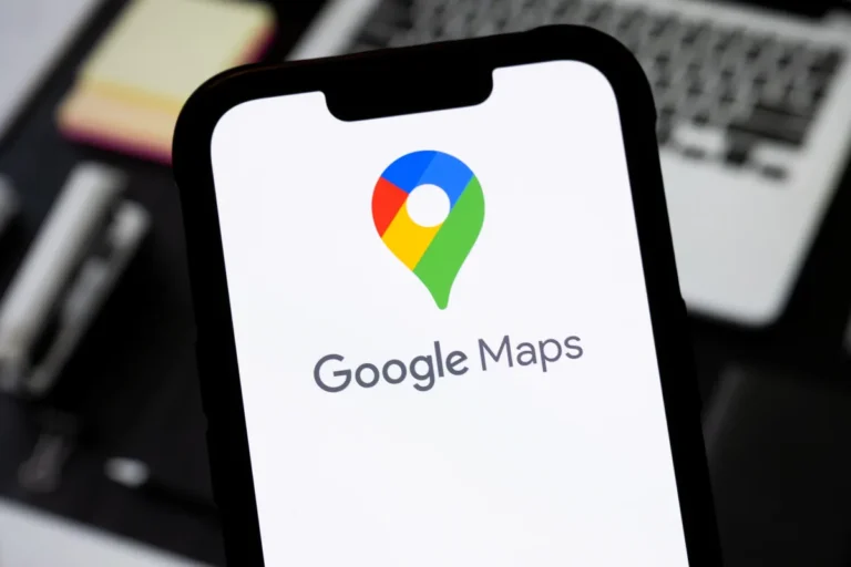 Smartphone displaying Google Maps logo and text, with a blurred laptop keyboard in the background.