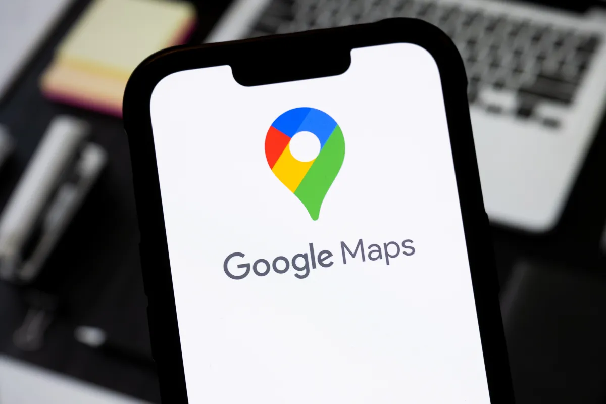 Smartphone displaying Google Maps logo and text, with a blurred laptop keyboard in the background.