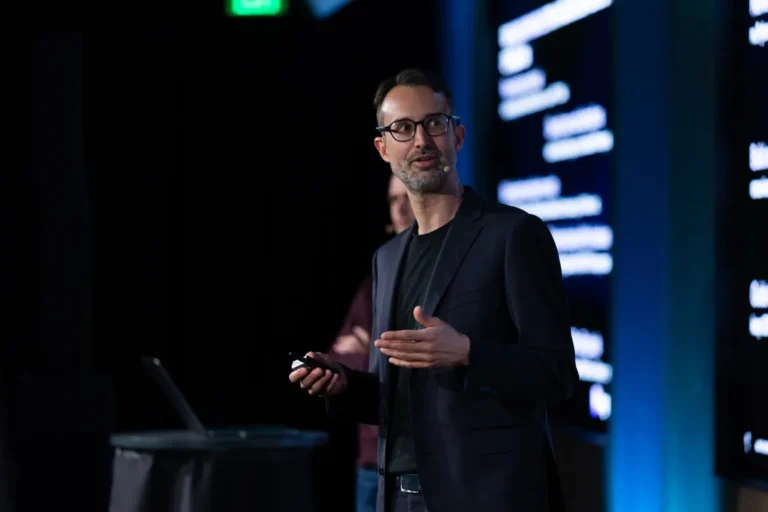 Man in a dark blazer speaking on stage with a headset, holding a remote, blurred text on screen in the background.