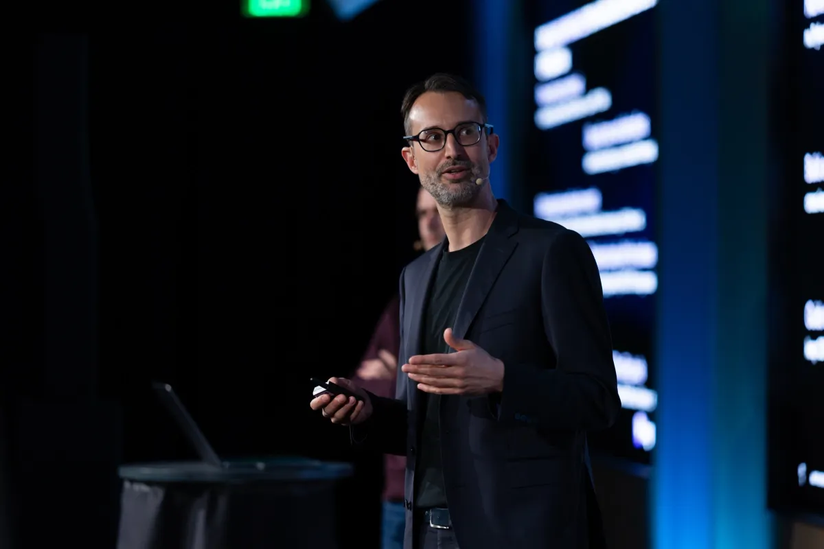Man in a dark blazer speaking on stage with a headset, holding a remote, blurred text on screen in the background.
