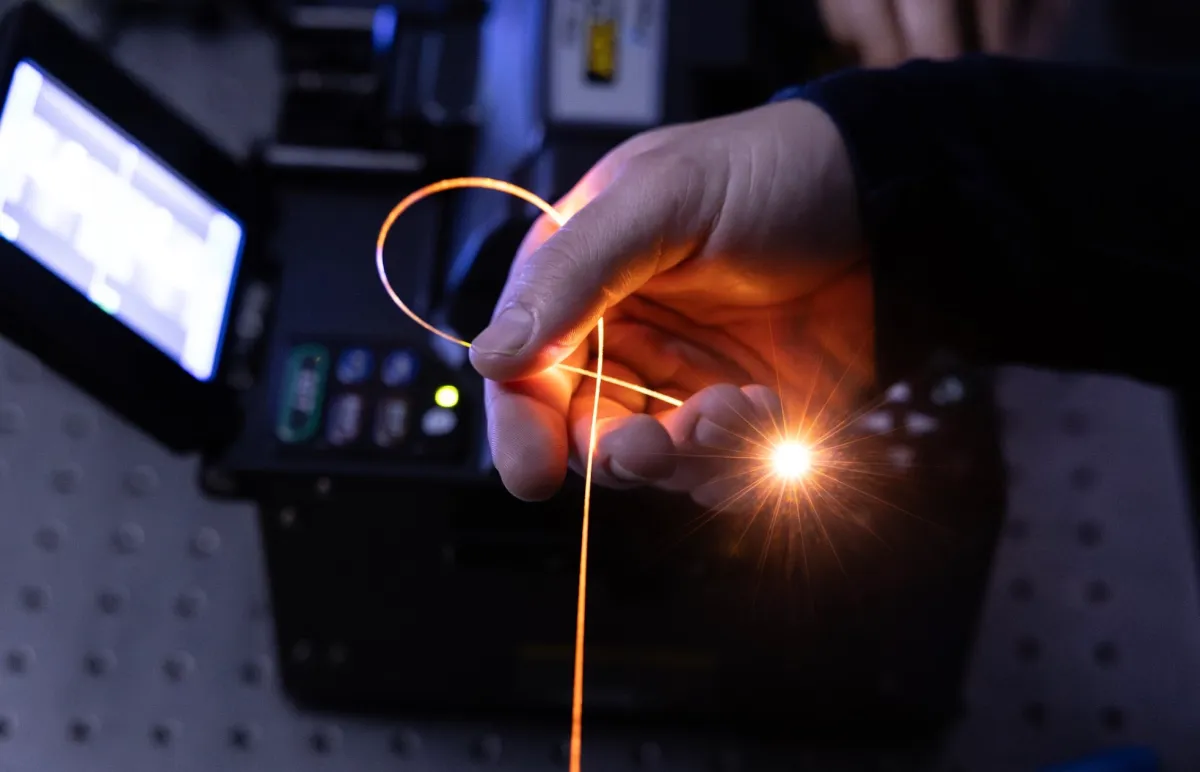 Hand holding glowing fiber optic cable looped, with equipment in the background.