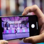 Hands holding a smartphone, capturing a photo of a colorful carnival game booth.