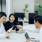 Three people in an office, two in white coats, discussing a handheld device at a table with laptops and plants in the back...