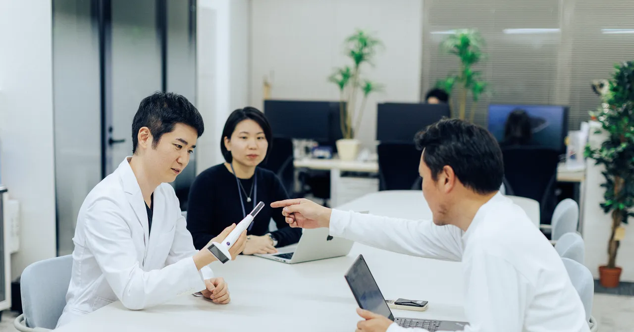 Three people in an office, two in white coats, discussing a handheld device at a table with laptops and plants in the back...