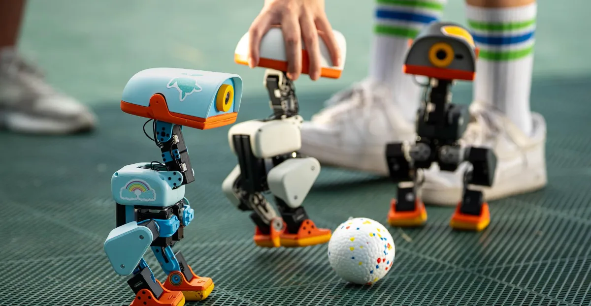 Small colorful robots on a green surface with a white ball; hand adjusting one robot, person wearing striped socks.